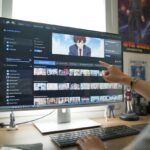 Um ambiente de trabalho moderno com um computador mostrando uma plataforma de streaming aberta com uma série de anime, com figuras e pôsteres de anime ao redor e uma mão apontando para a tela.