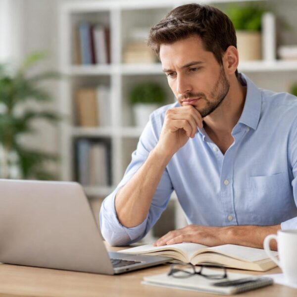Pessoa pensativa sentada em uma mesa moderna, olhando para um livro aberto ou laptop, em um escritório iluminado com livros e plantas ao fundo.