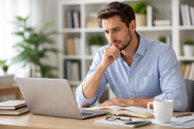 Pessoa pensativa sentada em uma mesa moderna, olhando para um livro aberto ou laptop, em um escritório iluminado com livros e plantas ao fundo.