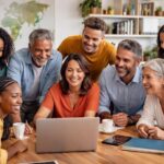 Grupo diversificado de pessoas brasileiras reunidas em um escritório moderno, discutindo em volta de uma mesa com laptops e cadernos.