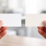 Duas mãos segurando dois pequenos cartões retangulares diferentes em um ambiente de escritório.